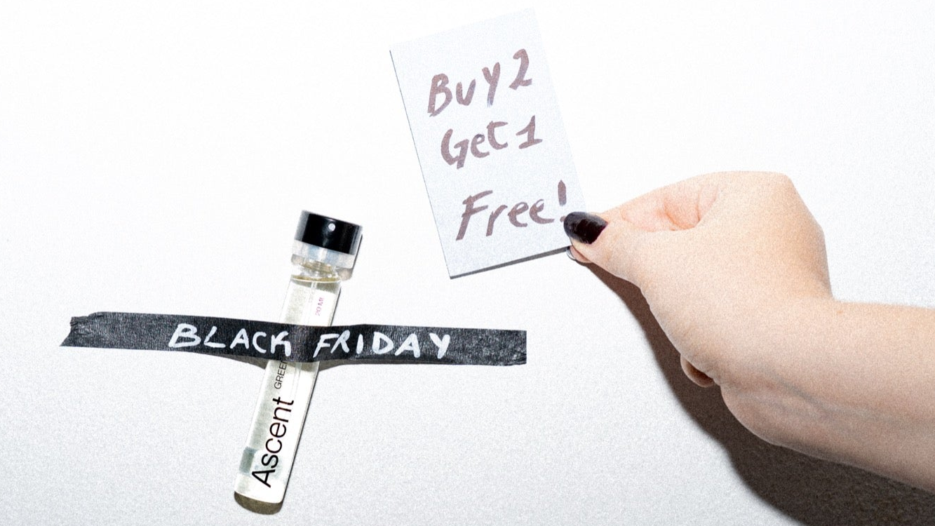 A small clear perfume sample vial with a black cap is taped to a white wall with black tape reading 'BLACK FRIDAY'. A hand with dark nail polish holds a small card that says 'Buy 2 Get 1 Free!'.