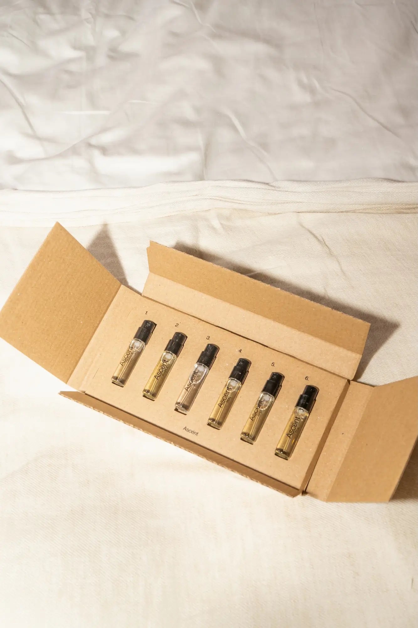 Open cardboard box containing six small perfume bottles on a white fabric background