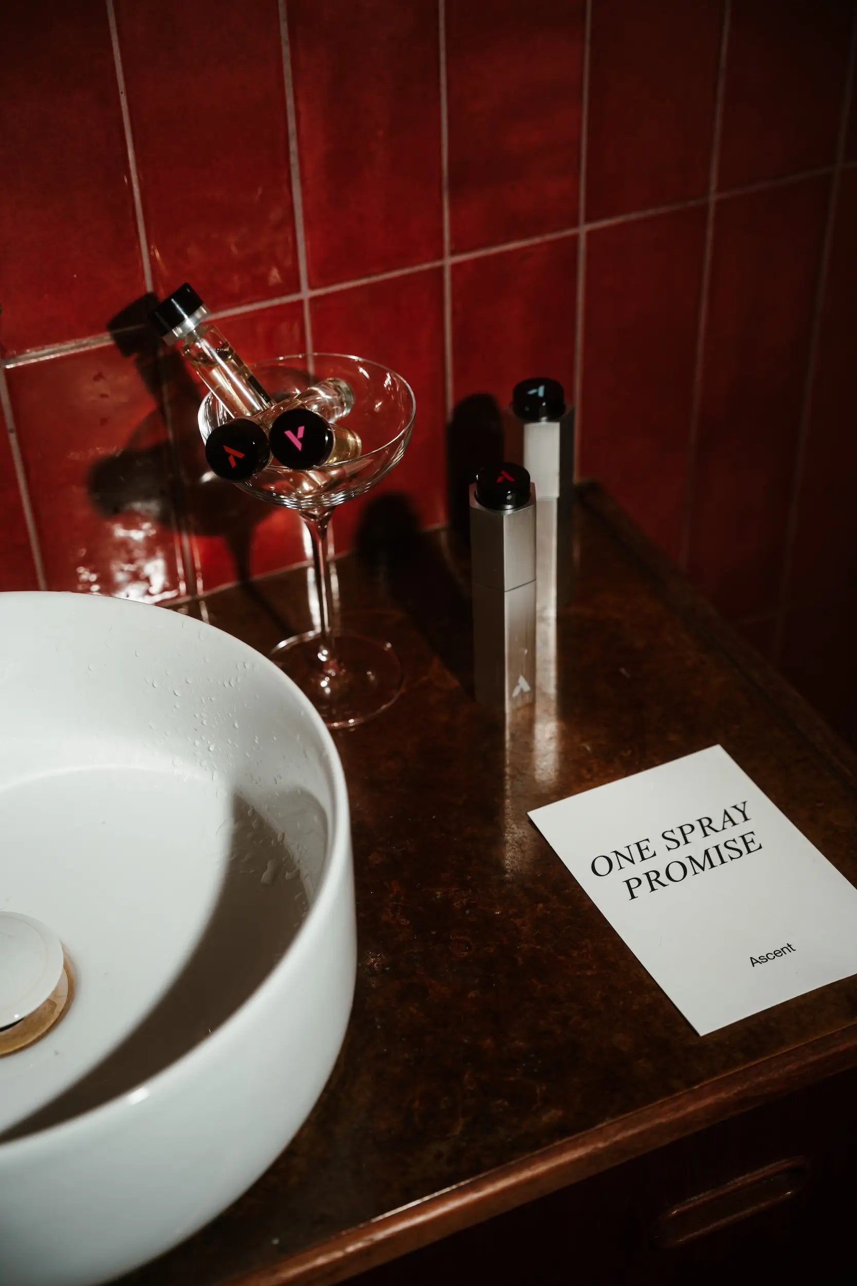 Bottle of perfume, glass, and a card on a counter with red tiled wall in the background